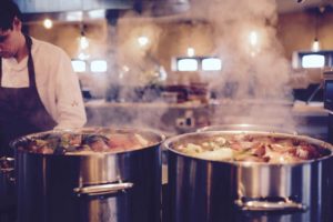 Male chef in a kitchen standing next to two huge pots cooking something